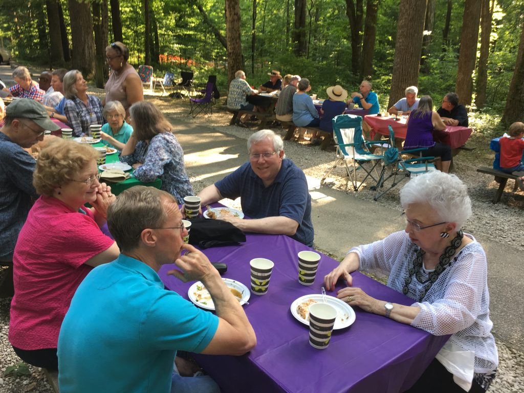 Group of people sitting at picnic tables outdoors, enjoying food and conversation at a picnic.