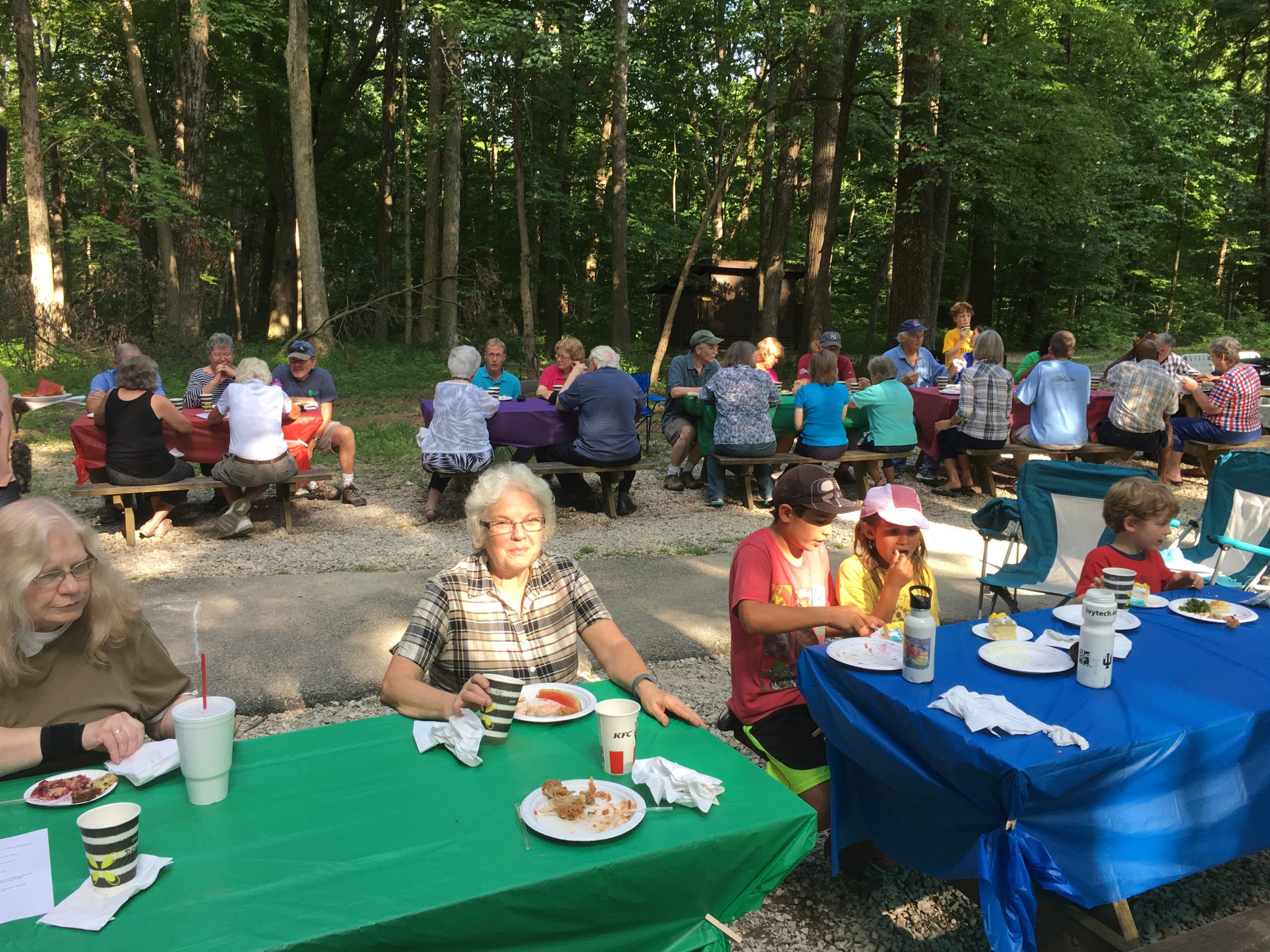 Large group of people sitting at multiple picnic tables outdoors, enjoying a meal together.