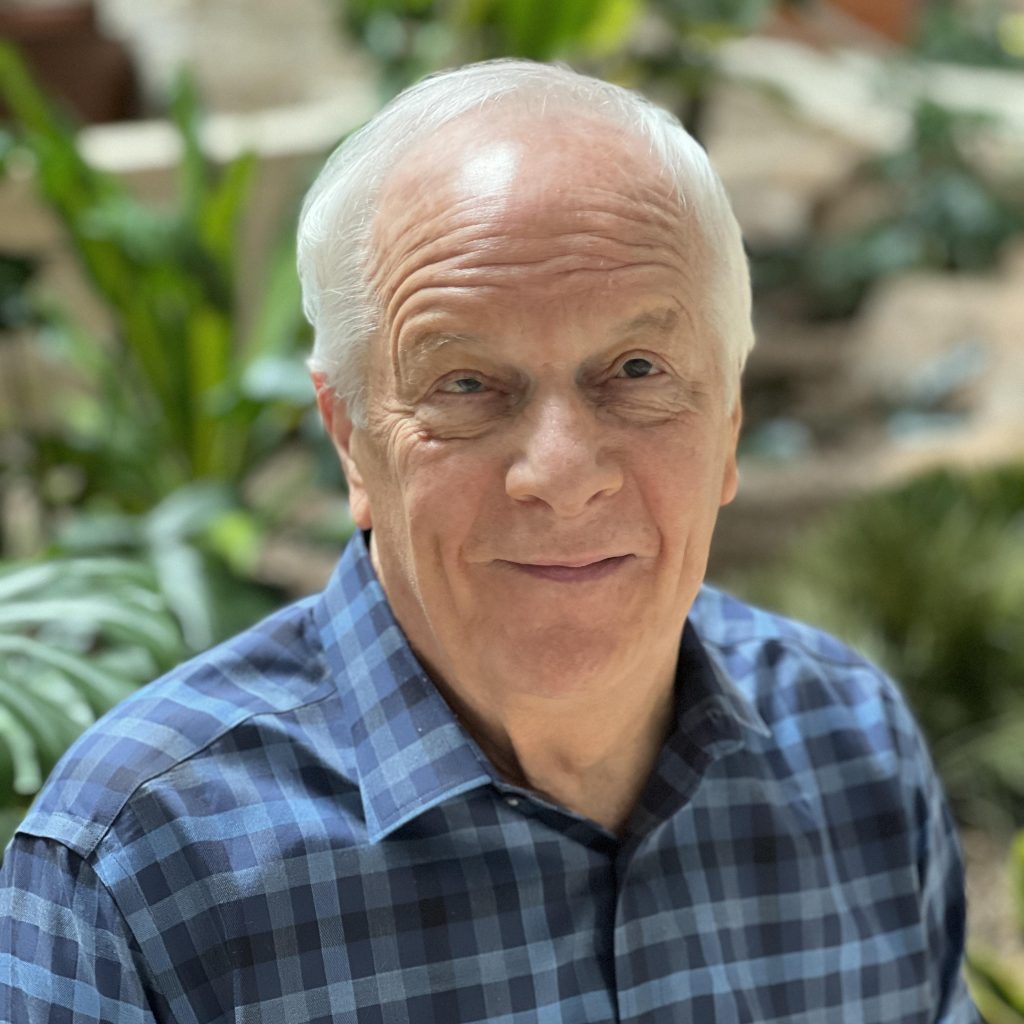 Ken Medema, a visually impaired musician and composer, smiling in an indoor setting with greenery in the background.